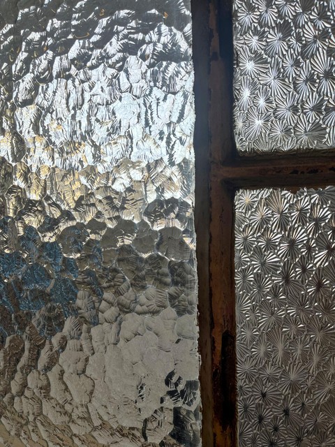 De foto toont een close-up van een raam met structuurglas. Op de linkerkant van de foto lijkt het patroon op ijsbloemen geribbeld met willekeurige, natuurlijke vorming, wat een ruw en onregelmatig oppervlak creëert. Aan de rechterkant is het patroon meer uniform en geometrisch, met herhalende vormen die lijken op sterren of bloemen. Het licht dat door het glas schijnt, benadrukt de texturen en geeft het een glinsterend effect.